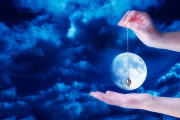Close up of woman's hand holding a pendulum over her palm. Cloudy sky and full moon at night in background.