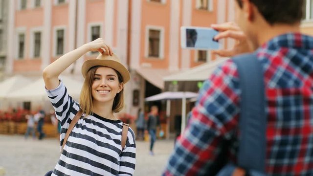 Young caucasian man taking photo of a young woman in hat and striped top using a cell phone. Photography from behind. Outdoors.