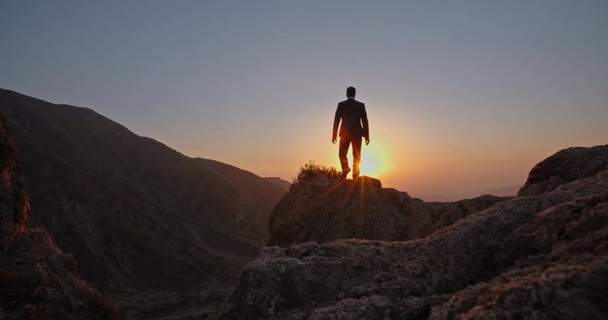 Young successful bsinessman wearing suit and tie standing on top of a mountain, raising his hands - way to success, on top of the world concept 4k