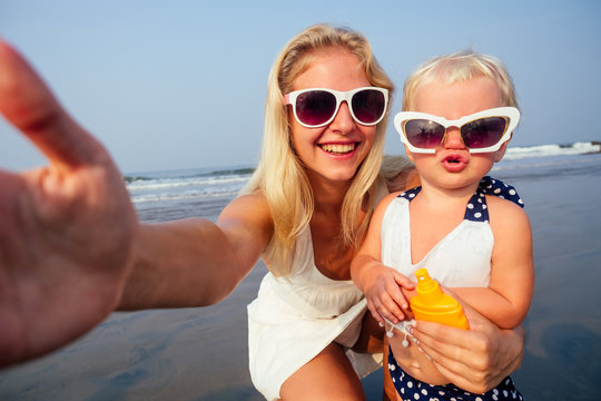 fair-haired mother in stylish glasses,white dress orange spray bottle SPF sunscreen with newborn baby one year old lips duck air kiss daughter taking portrait on the camera smartphone summer beach