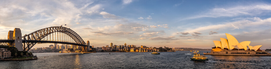 Panoramic view of Sydney harbor bridge and opera house with boats sailing in the bay and downtown in the background at sunset