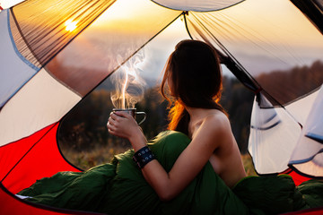 Naked woman sitting in tent in sleeping bag  and drinking coffee in the morning. A young female...