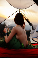 Naked woman sitting in tent in sleeping bag  and drinking coffee in the morning. A young female tourist drinks a hot drink and enjoy the scenery in the mountains at sunrise or sunset - 241826735