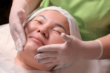 Fototapeta na wymiar Rejuvenating cosmetological procedure for a Muslim woman. The hands of a beautician in white gloves put the cream around the eyes on the face of an Asian patient. Face massage.