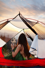 Naked woman sitting in tent in sleeping bag  and drinking coffee in the morning. A young female tourist drinks a hot drink and enjoy the scenery in the mountains at sunrise or sunset