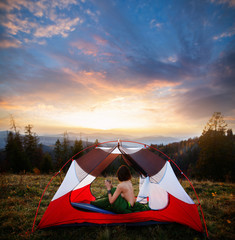 Rear view of naked woman sitting in tent in sleeping bag  and drinking coffee in the morning. A young female tourist enjoy the scenery in the mountains. Incredible cloudy sky at sunrise or sunset