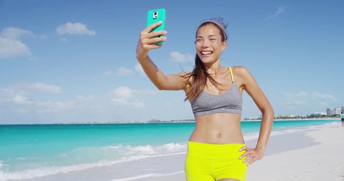 Sporty fresh woman taking selfie with phone laughing after running on beach. Girl taking fitness selfie after workout on summer vacation living healthy lifestyle having fun. RED EPIC SLOW MOTION