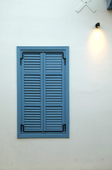 View of neat blue window with closed striped shutters on beige wall and a lamp in the upper right corner