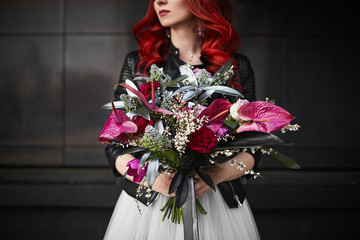 Fashionable and beautiful model girl with red hair and bright makeup, in a white wedding dress and in a leather jacket, with a big and luxury bouquet of exotic flowers in her hands, posing outdoors
