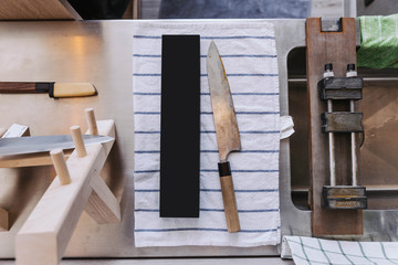 Dull and rustic Japanese kitchen knife waiting for sharpening on blue strip white fabric with sharpening equipment.