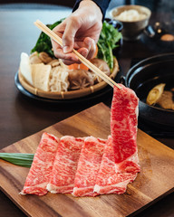 A hand holding Premium Rare Slices Wagyu A5 beef with high-marbled texture with chopsticks on square wooden plate served for Sukiyaki and Shabu.