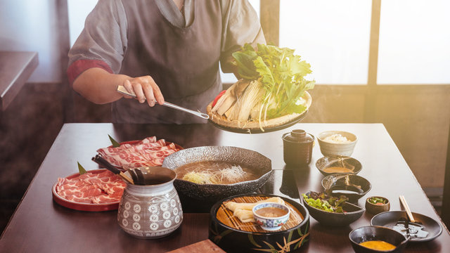 A woman holding vegetables into hot pot by tongs with Wagyu A5 beef and sliced Kurobuta in Shabu clear soup with stream.
