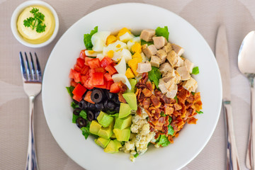 Cobb salad on white plate, with bacon, blue cheese, grilled chicken, cubed cut tomatoes and avocados, olives, and chopped lecttuce