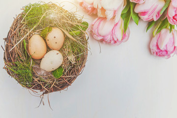 Fototapeta na wymiar Spring greeting card. Easter eggs in nest with moss and pink fresh tulip flowers bouquet on rustic white wooden background. Easter concept. Flat lay top view copy space. Spring flowers tulips