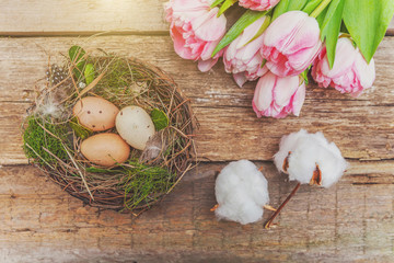Spring greeting card. Easter eggs in nest with moss and pink fresh tulip flowers bouquet on rustic shabby wooden background. Easter concept. Flat lay top view copy space. Spring flowers tulips