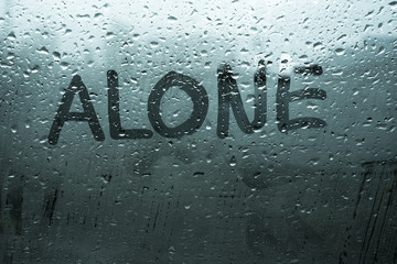 Handwritten word alone on fog up glass window with rain drops in cold dark tones, illustrates sad emotions, unhappy feelings, depression, loneliness and sadness mood