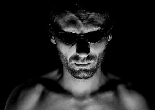 Portrait of unshaven adult caucasian man in swimming glasses. He smiles and looks straight to watcher. Black and white shot, low-key lighting. Isolated on black.
