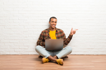 African american man sitting on the floor with his laptop pointing finger to the side in lateral...