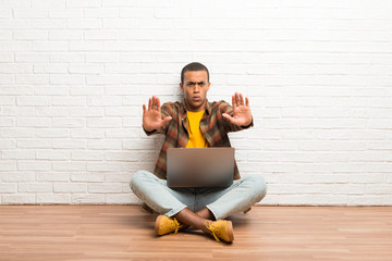 African american man sitting on the floor with his laptop making stop gesture for disappointed with an opinion