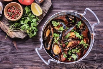 Delicious freshly cooked black mussels in tomato sauce in a vintage saucepan next to the ingredients on a wooden background, top view