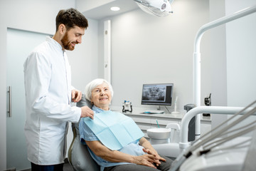 Handsome dentist fixing napkin on the elderly woman patient preparing for the procedure in the dental office