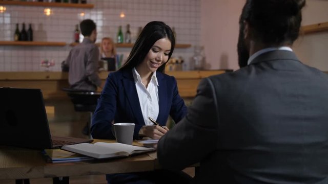Positive happy mixed race woman singing mortgage agreement with real estate agent. Happy female owner and male real estate broker handshaking after signing a contract for house investment at cafe.