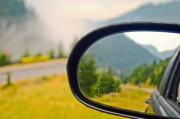 rear view mirror in focus, with a soft blurry  reflection of a mountain road against backdrop of the blue cloudy sky