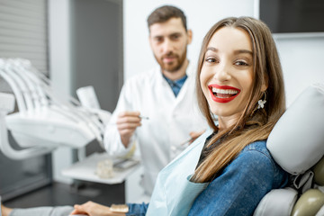 Obraz premium Portrait of a young smiling woman with healthy smile sitting in the dental office with male dentist on the background