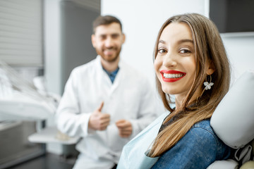 Portrait of a young smiling woman with healthy smile sitting in the dental office with male dentist on the background