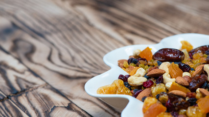 Dried fruits in white plate on wooden table, copy space for text. Mix of different varieties of nuts and berries, vitamins