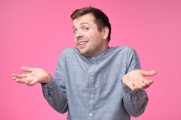Uncertain young stylish man wears blue shirt, shrugs shoulders being puzzled or confused.