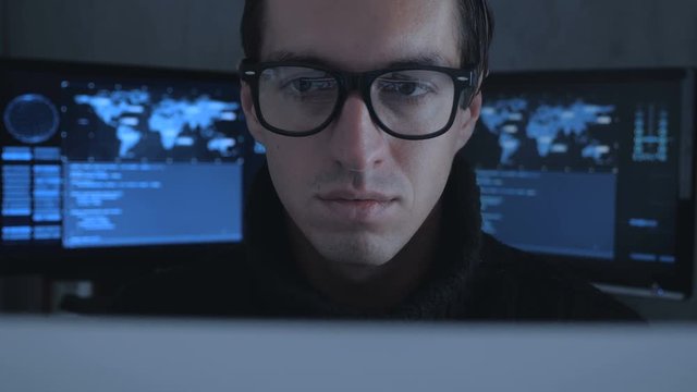 Hacker programmer in glasses is working on computer while blue code characters reflect on his face in cyber security center filled with display screens.
