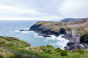 dark, black steep cliffs on the sea, where only the grass grows; blue-green sea with white waves on rocky shores; sea landscape with rocks and sea