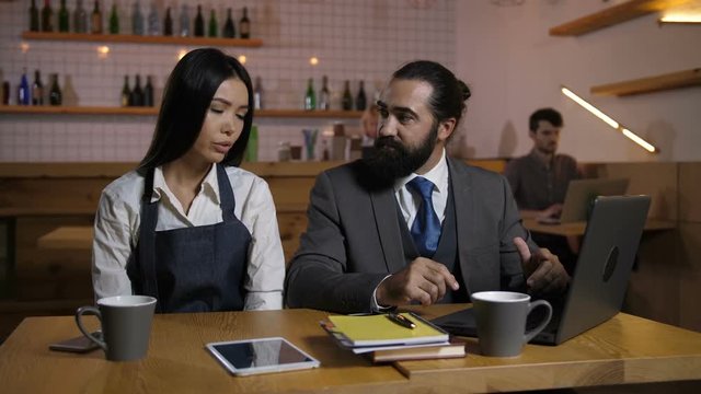 Irritated bearded male manager scolding, criticizing stressed female employee for incompetence at cafe. Sad beautiful employee in apron receiving fair reprimand from restaurant owner indoors.