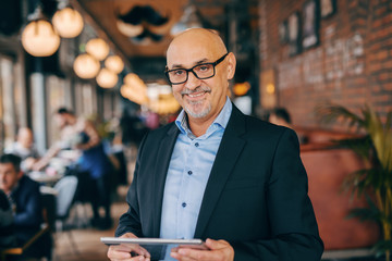 Smiling bearded senior in suit using tablet while standing in cafeteria.