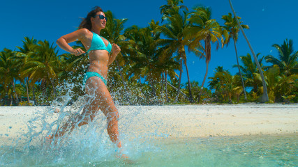 LOW ANGLE: Crystal clear ocean water splashing around girl running along shore.