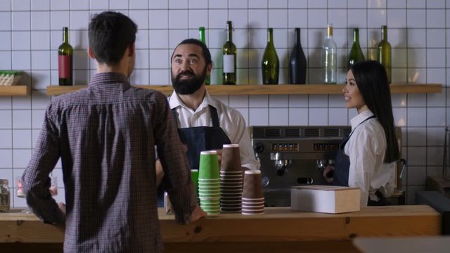 Positive bearded male barista in good mood communicating with client, offering best coffee drinks while helping customer to make choice with joyful mixed race waitress working at coffee shop counter
