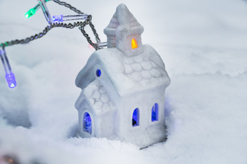 Naklejka premium A beautiful fabulous little white house with yellow and blue lights in the windows and garlands of burning colorful lamps on a white snow background in the holiday evening