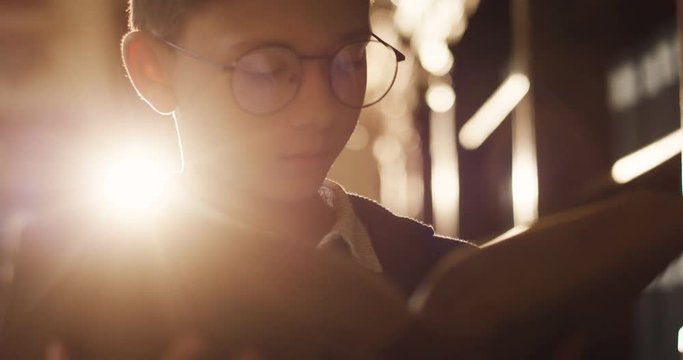 Close up of the teen boy in glasses reading a book while standing in the dark library in sunlight.