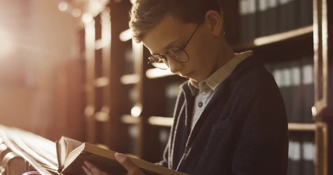 Portrait shot of the cute blond schoolboy in glasses reading a book attentively while standing in the library passage in sunlight.