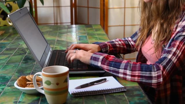 Woman at the table working with laptop. Girl wearing a checkered shirt is typing on her black keyboard with coffee and cookies by her side. Fixed angle scene from the left side