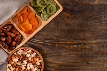Dates, dried apricots and kiwis in a Compartmental dish and assortment of nuts in wooden bowl on a dark wooden table.