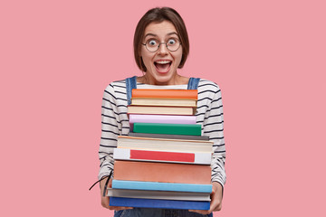 Waist up shot of joyful student in round spectacles, holds pile of books, prepares for seminar or writing report, opens mouth widely, isolated over pink background. Intelligent female studies indoor
