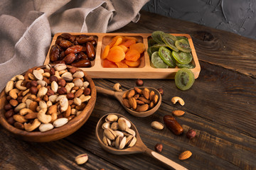 Dates, dried apricots and kiwis in a Compartmental dish and assortment of nuts in wooden bowl on a dark wooden table.