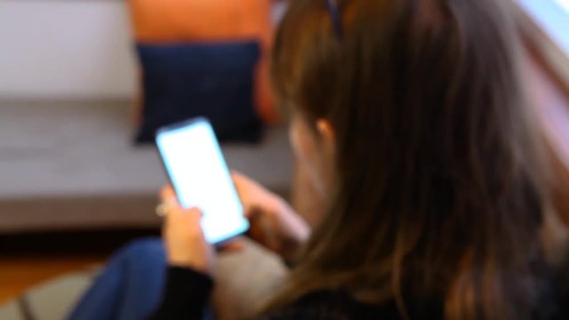 Girl using mobile phone in a living room. Young woman with wooly grey shirt is scrolling on her phone at home. Fixed scene from the back that goes in and out of focus