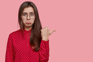 Unhappy gorgeous woman wants new expensive dress from shop, frowns and points aside, expresses displeasure, has no money for buying it, wears red polka dot shirt, models over pink studio wall