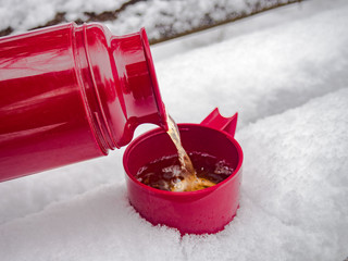 Pouring hot tea from thermos into plastic cup standing on bench covered with snow - obrazy, fototapety, plakaty