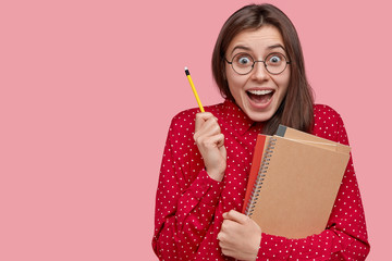 Cute young woman with joyful expression carries pencil and notepad, opens mouth widely, makes notes in notebook, stands over pink background with blank space on right side for your promotion. - Powered by Adobe