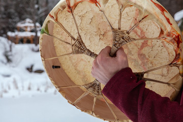 Man is playing native sacred drum. Young man wearing a red shirt is pictured from the back is playing his sacred drum with a drumstick. Pictured from the back, outside in winter