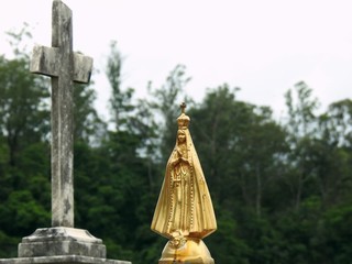 Scene in a graveyard: golden statue of Our Lady of Aparecida, widely venerated by Brazilian Catholics, next to an unfocused stone religious cross. In the blurred background, vegetation.